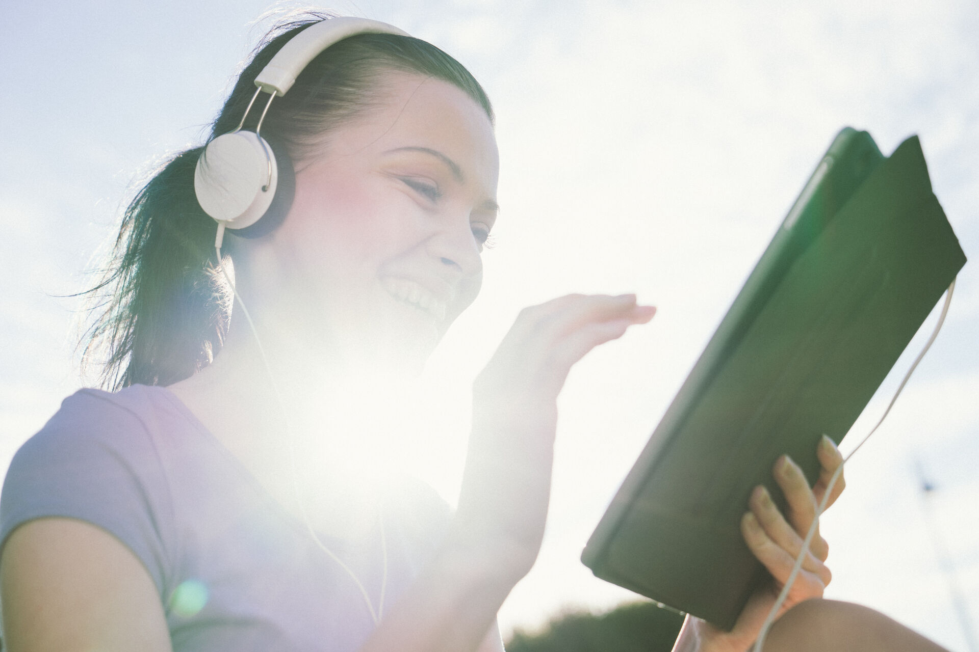Junge Frau guckt mit Kopfhörern im Sonnenschein auf ein Tablet